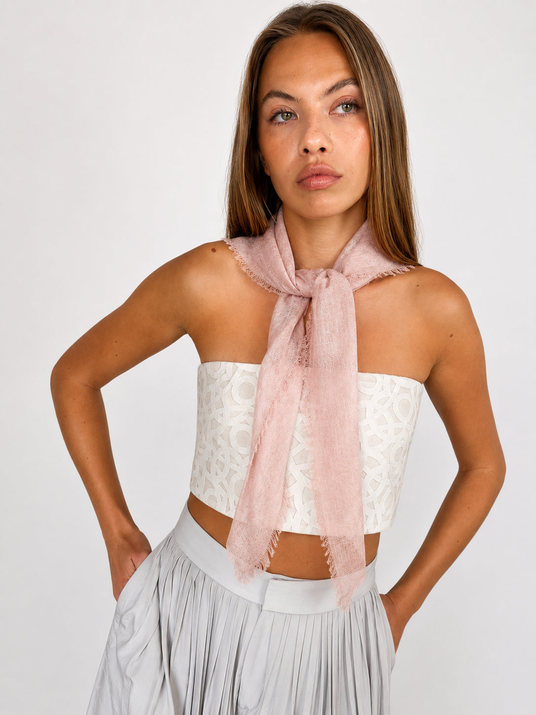 A woman modeling a light pink sheer scarf tied around her neck, paired with a white lace strapless top and a light gray pleated skirt, against a plain light gray studio background.