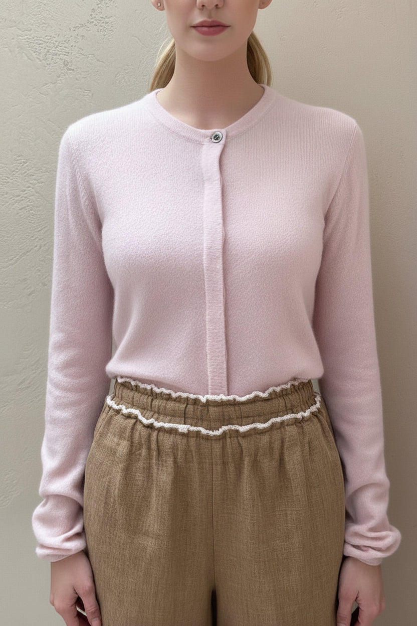 A woman wearing a pale pink, long sleeve, round neck cardigan with hidden buttons, paired with beige trousers.