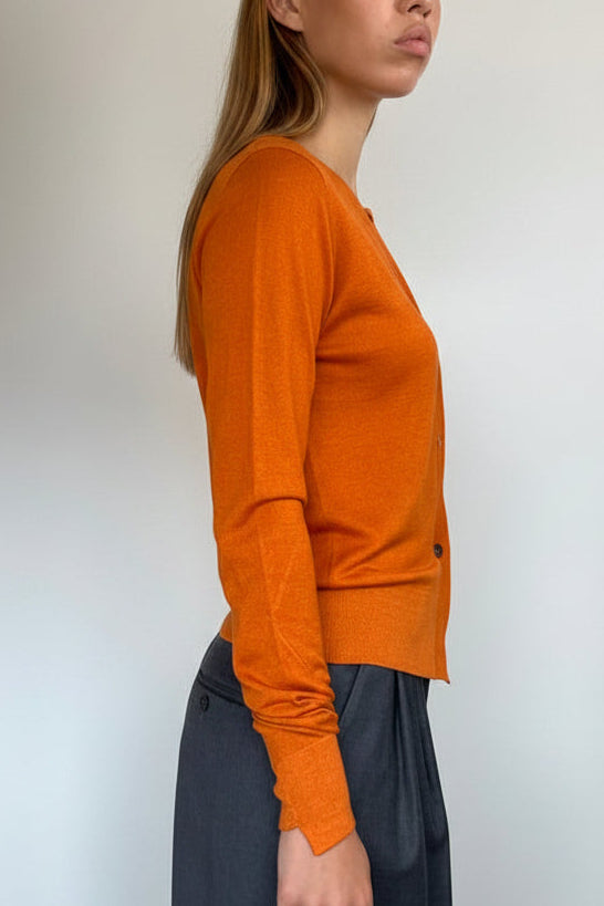 A woman wearing a fitted orange long-sleeve knit top and a dark gray skirt, standing in profile against a plain white wall.