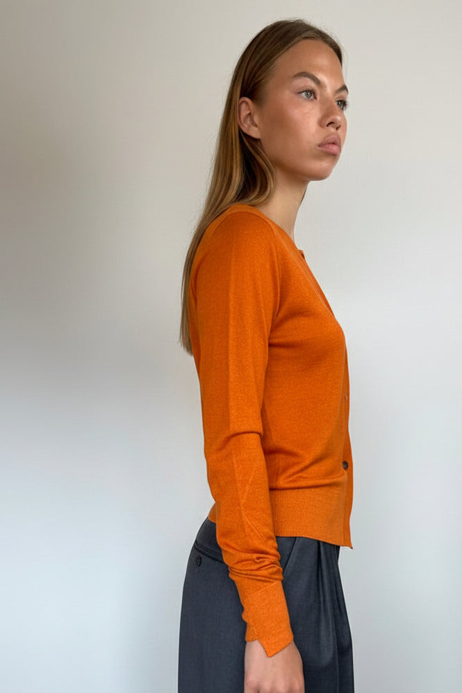 A woman wearing a fitted orange long-sleeve knit top and a dark gray skirt, standing in profile against a plain white wall.