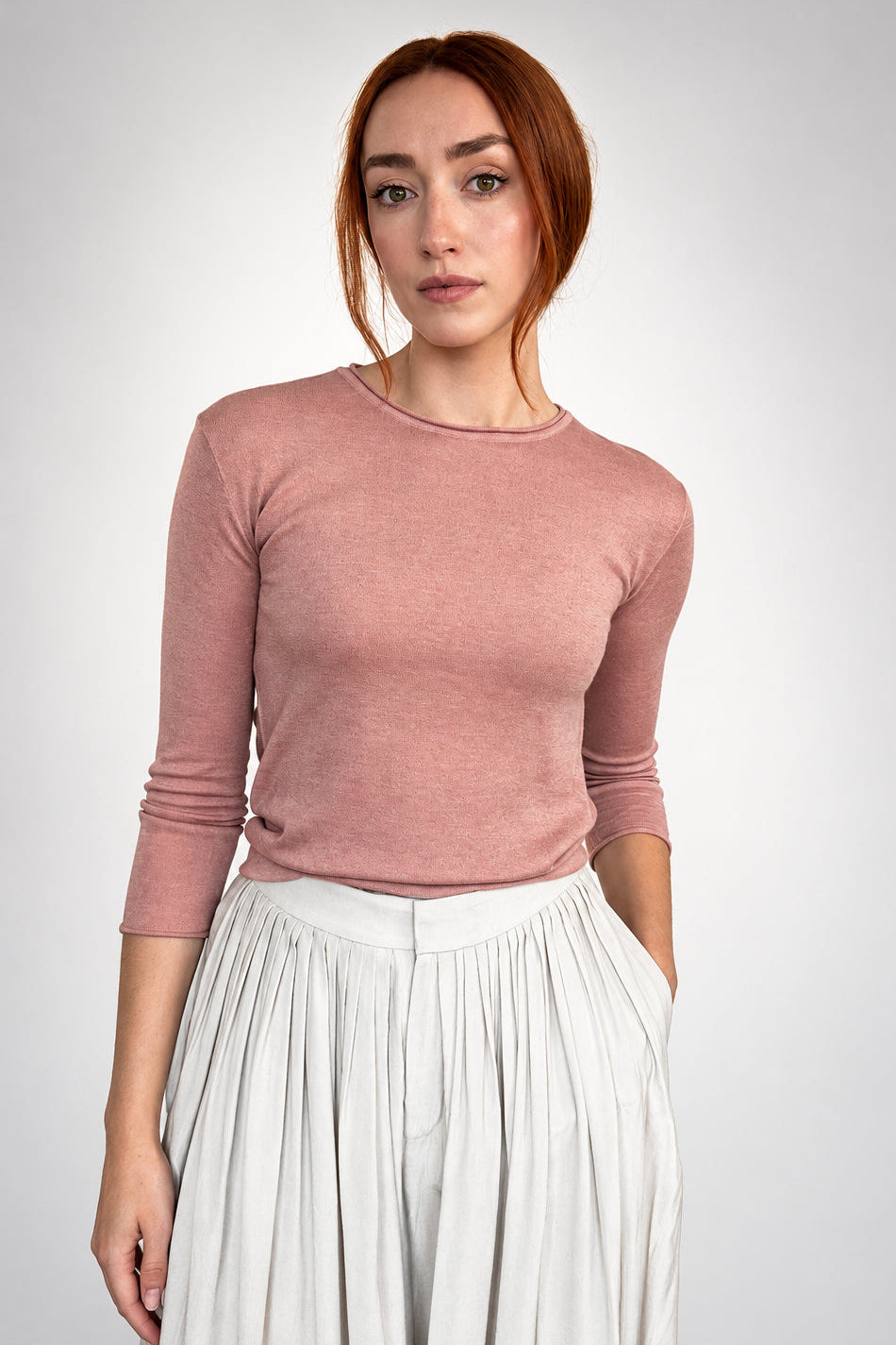 A woman with red hair stands against a plain light gray wall, wearing a fitted dusty rose long-sleeve top and a light gray pleated skirt.