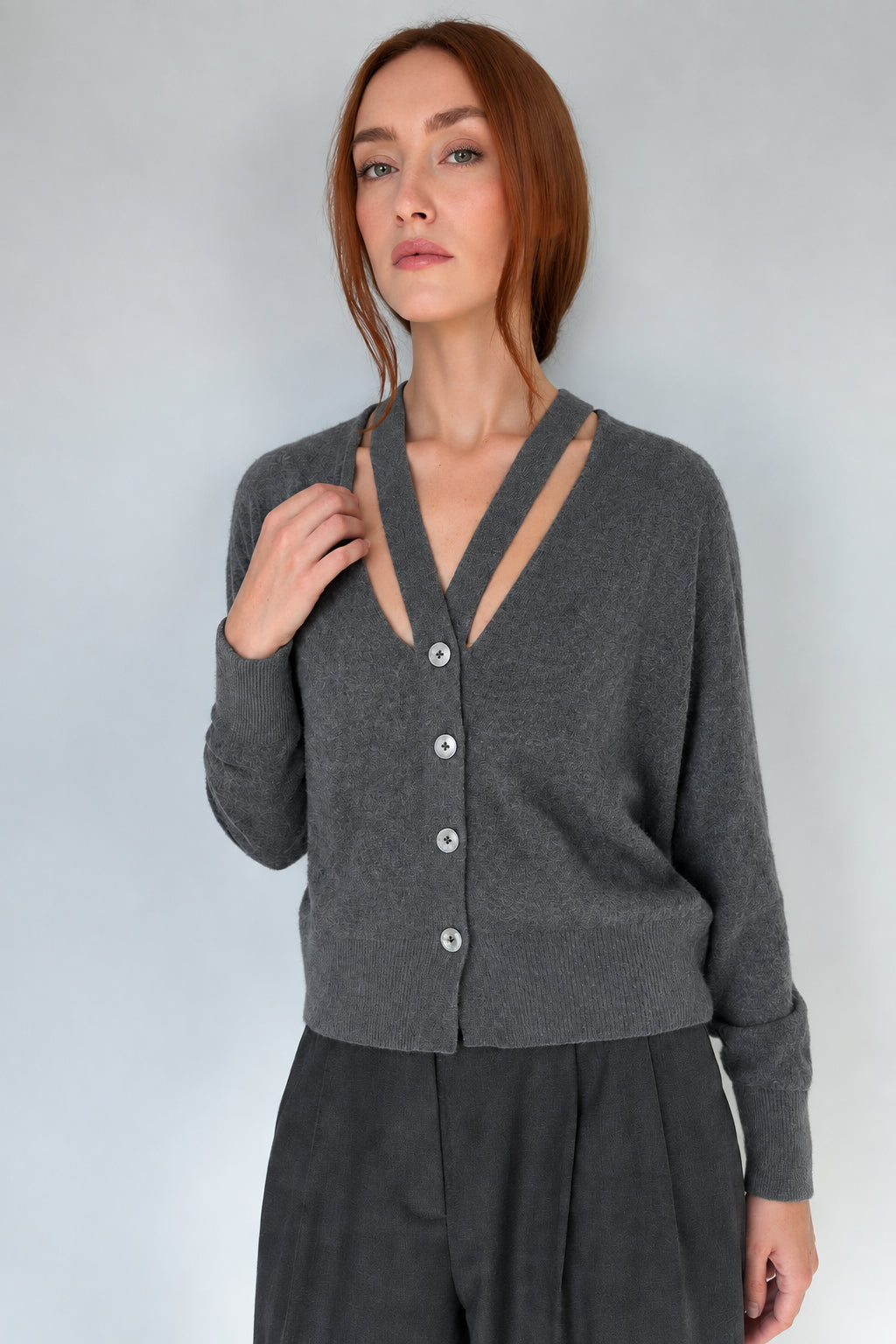 A woman with red hair wearing a dark grey melange  button-up cardigan with a split V-neckline and gray trousers, standing against a plain light gray wall.