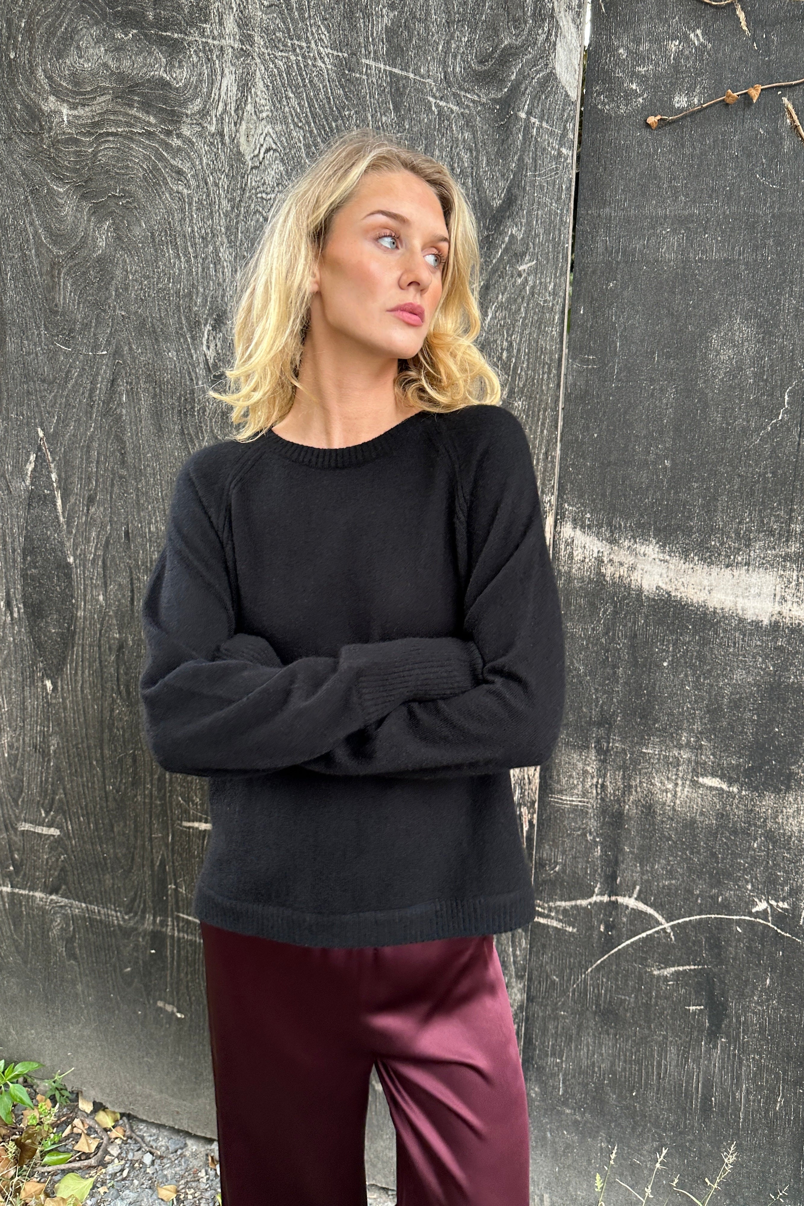 Woman wearing a black cashmere sweater and burgundy pants standing against a textured wall.