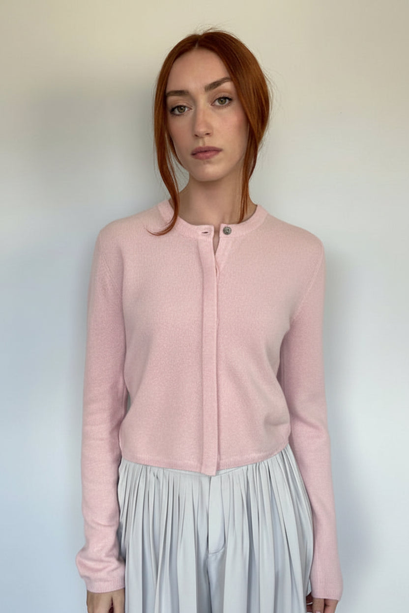 A woman with red hair stands against a plain off-white wall wearing a light pink long-sleeve knit cardigan with a buttoned round neckline over a light gray pleated skirt.