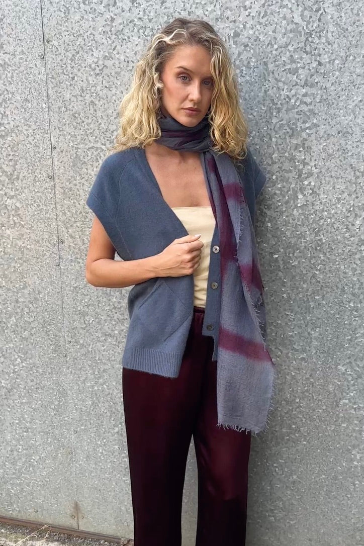 model wearing a blue  cashmere sleeveless cardigan  and plaid scarf against a textured wall.
