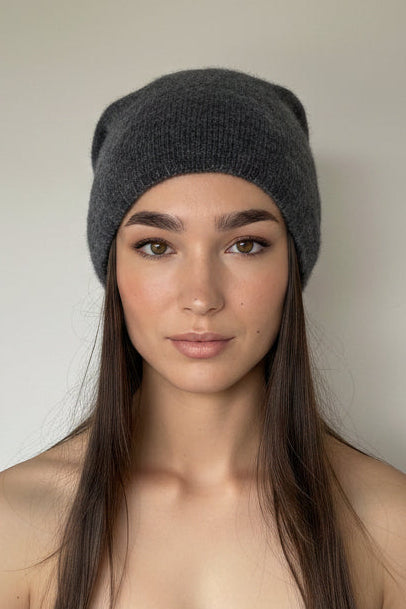 A woman with long dark brown hair wearing a dark gray knit beanie and a strapless taupe top, standing against a plain off-white wall.