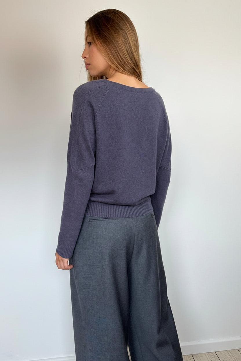 A woman wearing a slate purple knit sweater and dark gray wide-leg trousers, shown from a side-back angle against a plain white wall.