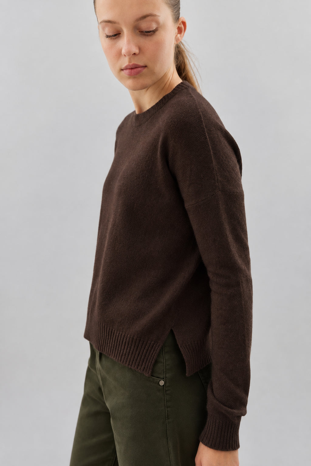A woman wearing a dark brown crewneck knit sweater and dark olive green pants, shown from a side profile against a plain light gray background.