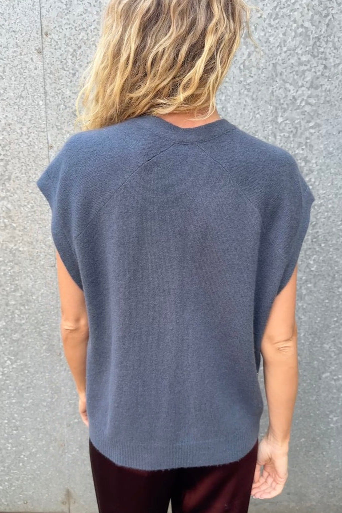 model wearing a blue  cashmere sleeveless cardigan  and plaid scarf against a textured wall. back view