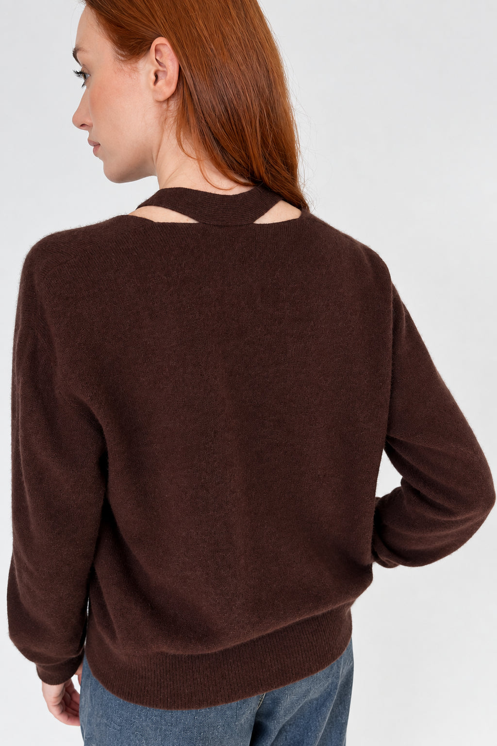 A woman with red hair is shown from the back wearing a brown knit sweater with a cutout at the neck and light gray pants, standing against a plain light gray wall.