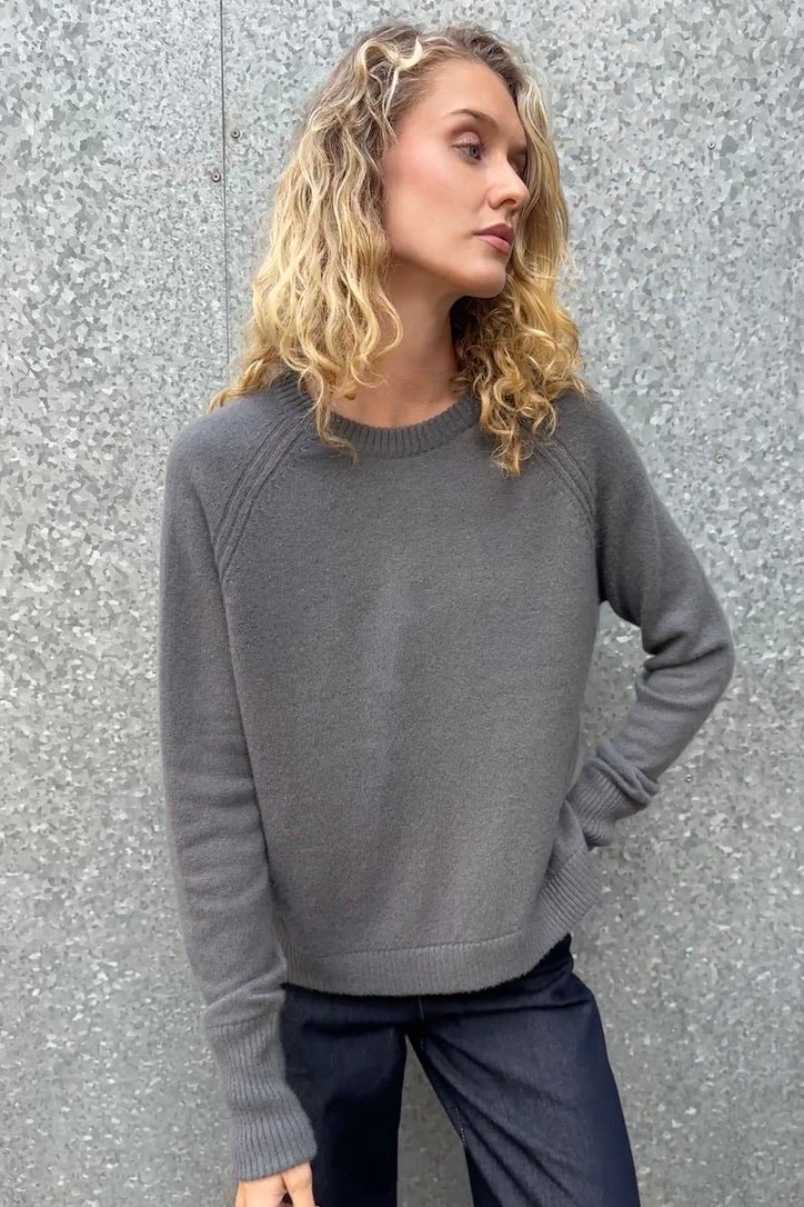 Woman wearing a hethna grey gray cashmere 
sweater against a textured gray wall
