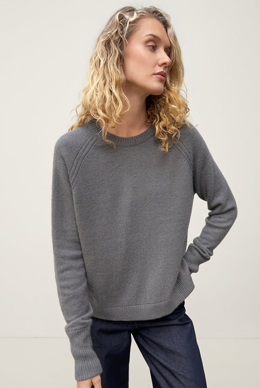 Woman wearing a hethna grey gray cashmere 
sweater against a textured gray wall