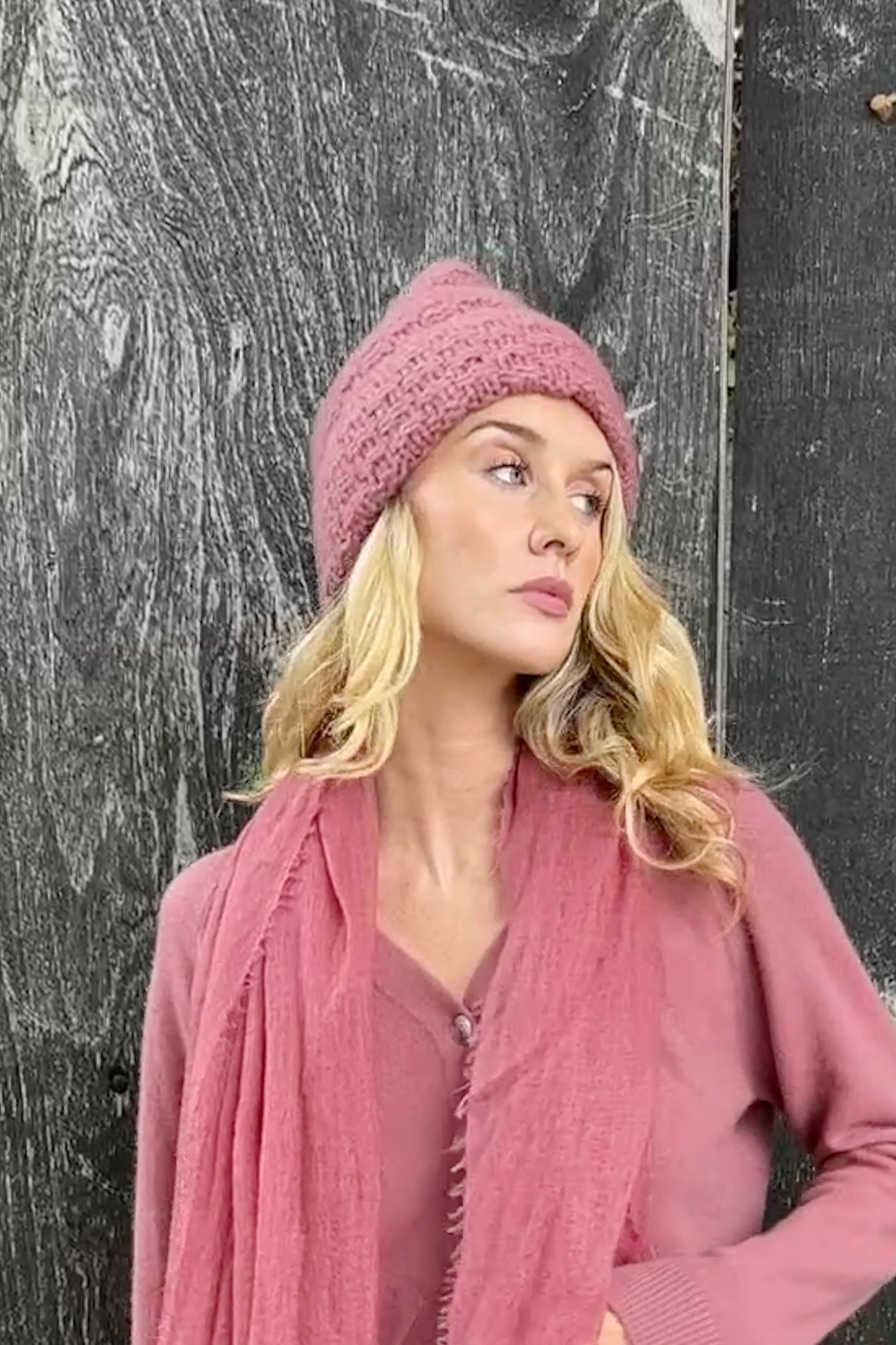 Woman wearing a pink knit beanie , margo beanie hat in brown pink cardigan against a textured black background