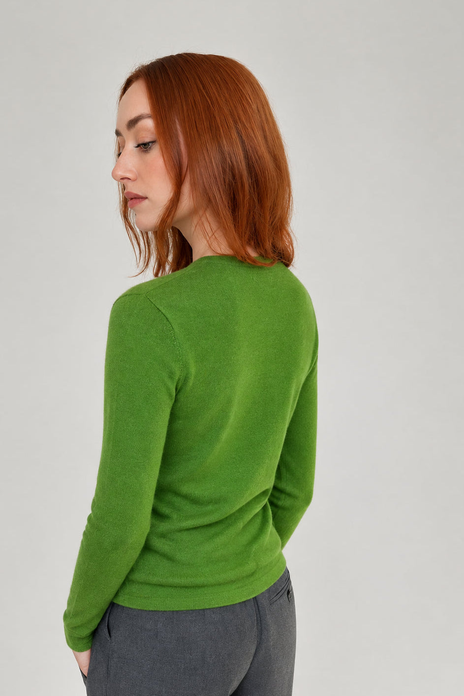 A woman with red hair is shown from a side-back angle wearing a bright green long-sleeve knit top and gray trousers against a plain light gray studio background.