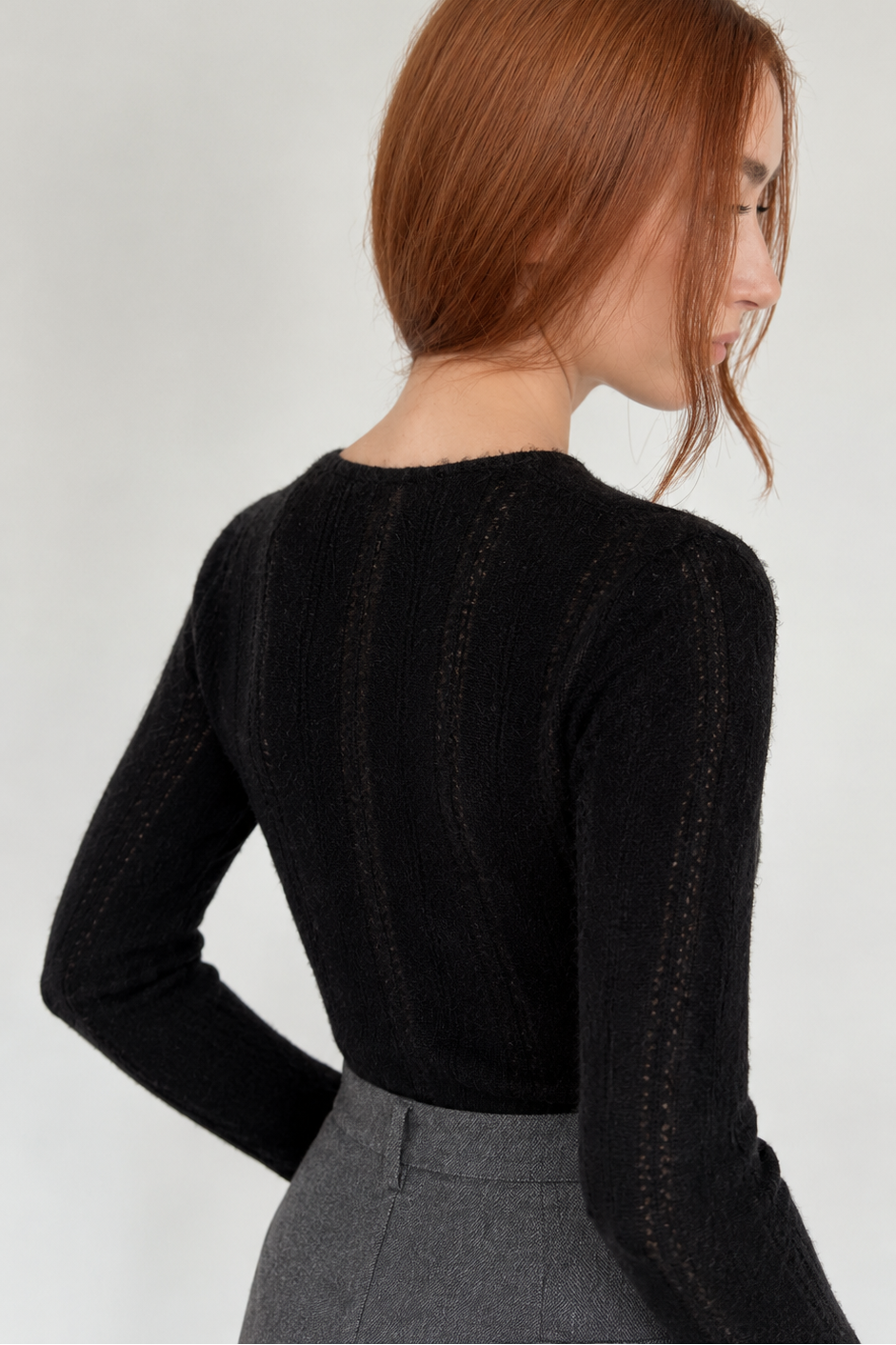 A woman with red hair is shown from the back wearing a black ribbed knit long-sleeve top and gray trousers against a light gray background.