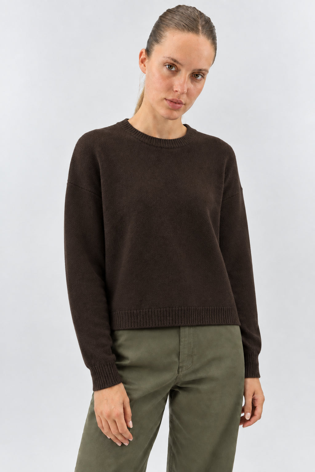 A woman wearing a dark brown crewneck knit sweater and dark olive pants, standing against a light gray studio background.