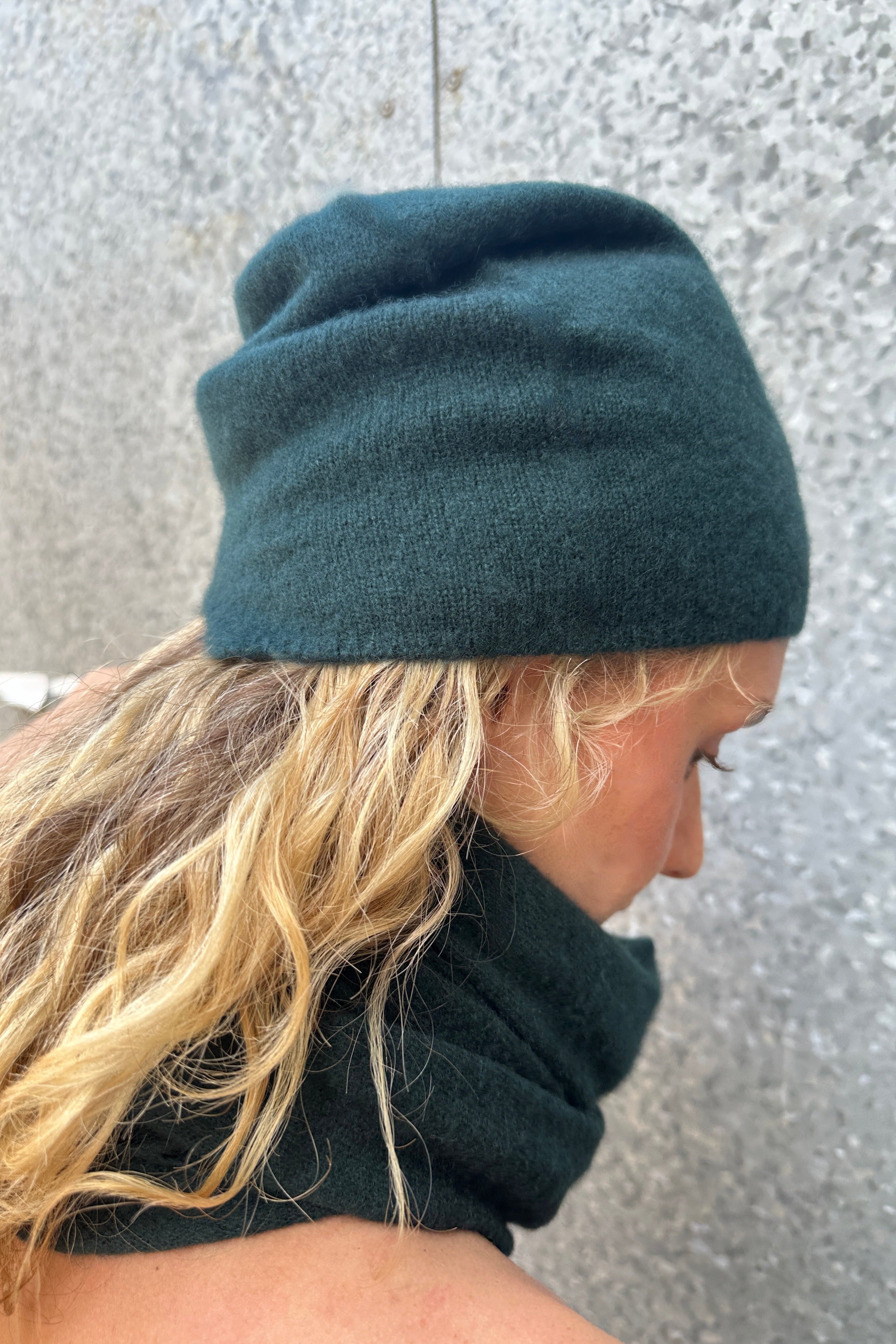 Model wearing a dark  teal blue beanie and matching scarf against a textured wall.