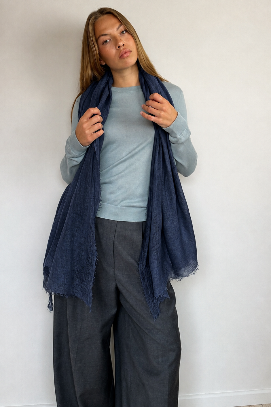 A woman modeling a dark navy blue scarf draped over her shoulders, paired with a light blue long-sleeve top and dark gray wide-leg pants, standing against a plain off-white wall.