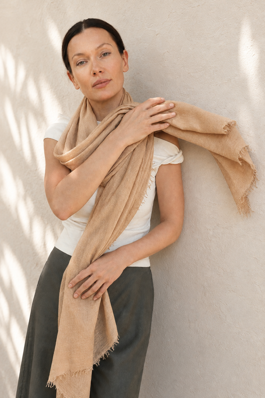 A woman wearing a light beige fringed scarf draped over her shoulders, standing against a textured beige wall with soft leaf shadows.