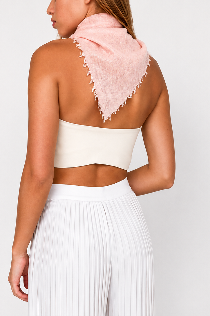 A woman shown from the back wearing a light pink fringed scarf draped over her shoulders, a white bandeau-style crop top, and white pleated pants, against a plain light gray studio background.