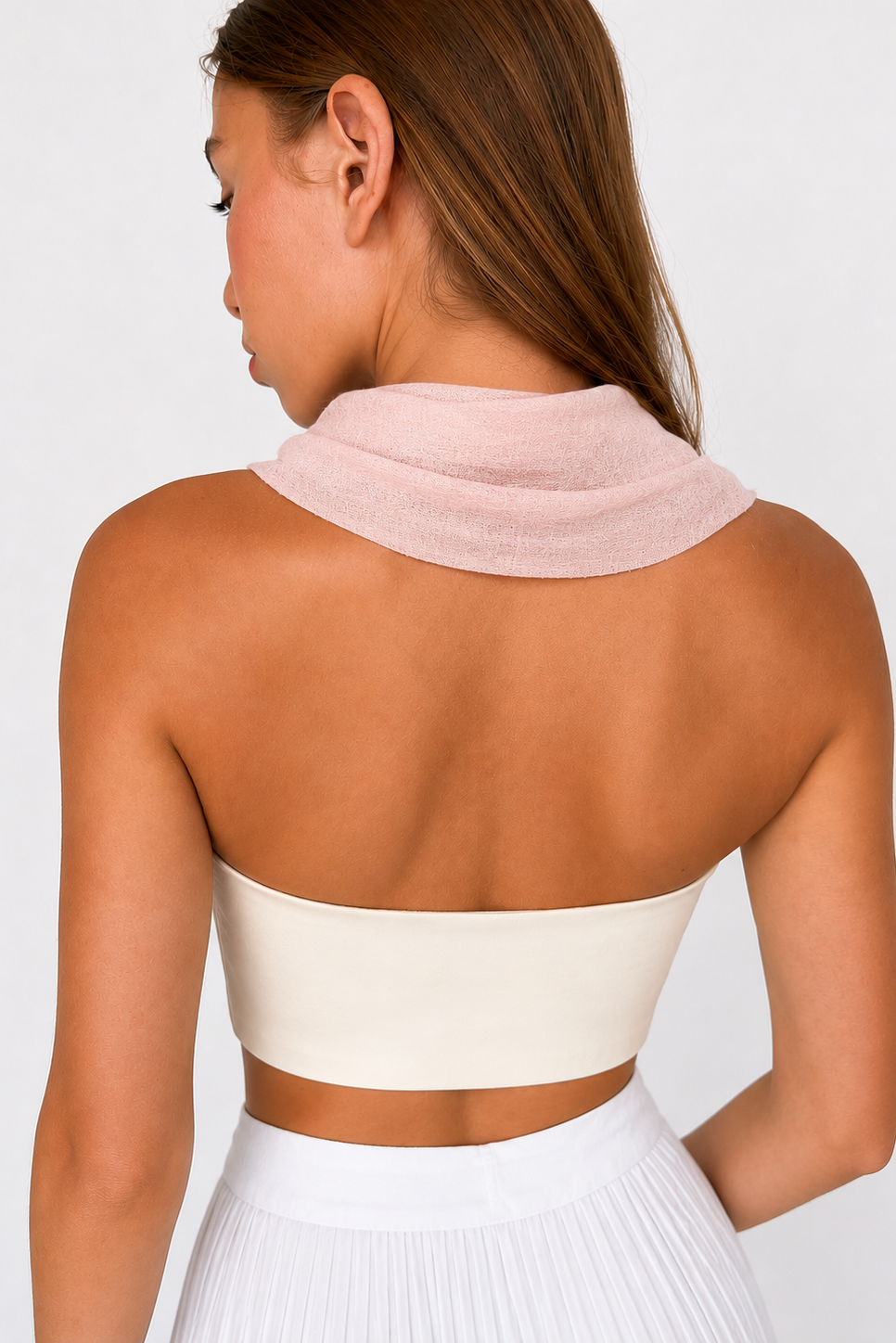 A woman shown from the back wearing a pale pink knit cowl neck scarf over her shoulders, paired with a white strapless tube top and white pleated skirt, against a light gray studio background.