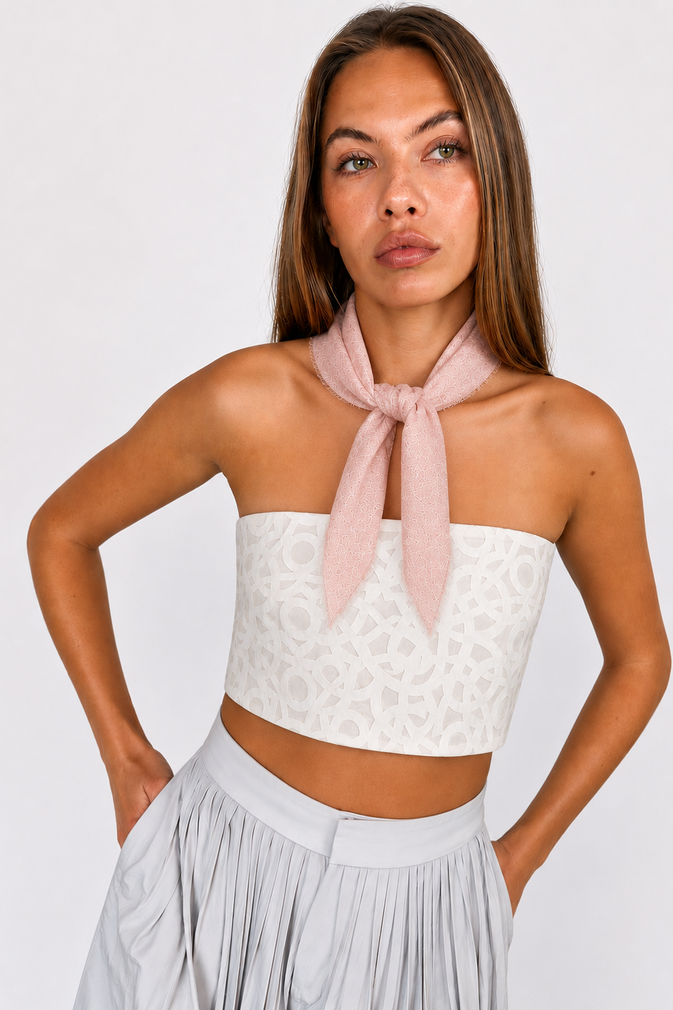 A woman modeling a white strapless tube top with a tonal geometric pattern, styled with a dusty pink neck scarf and a light gray pleated skirt against a plain light gray background.