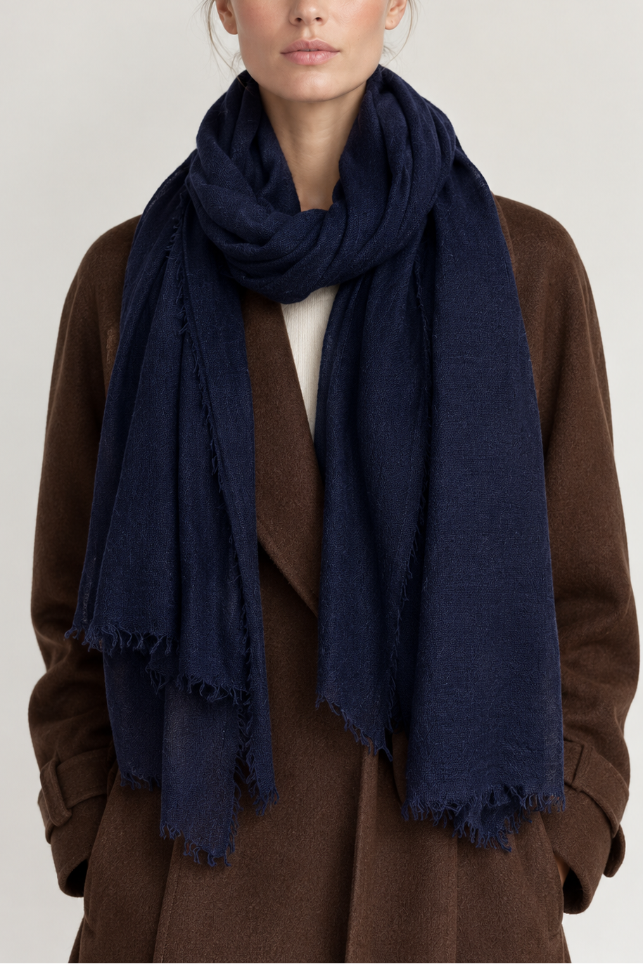 A woman wearing a dark navy blue scarf with fringed edges, styled over a brown coat and white top, photographed against a neutral light gray background.