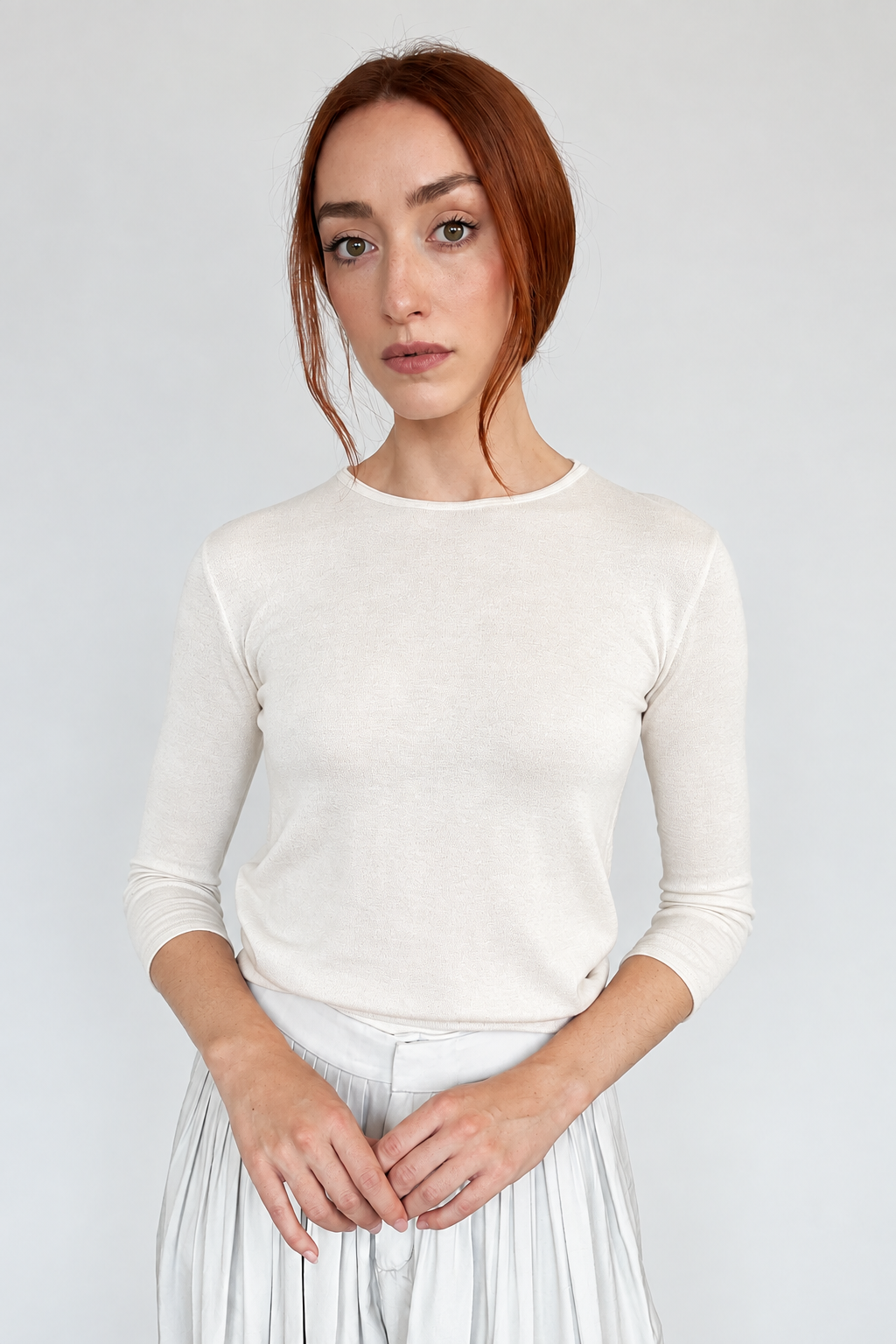 A woman with red hair wearing a light cream-colored long-sleeve crewneck knit top and a white pleated skirt, standing against a plain light gray wall.