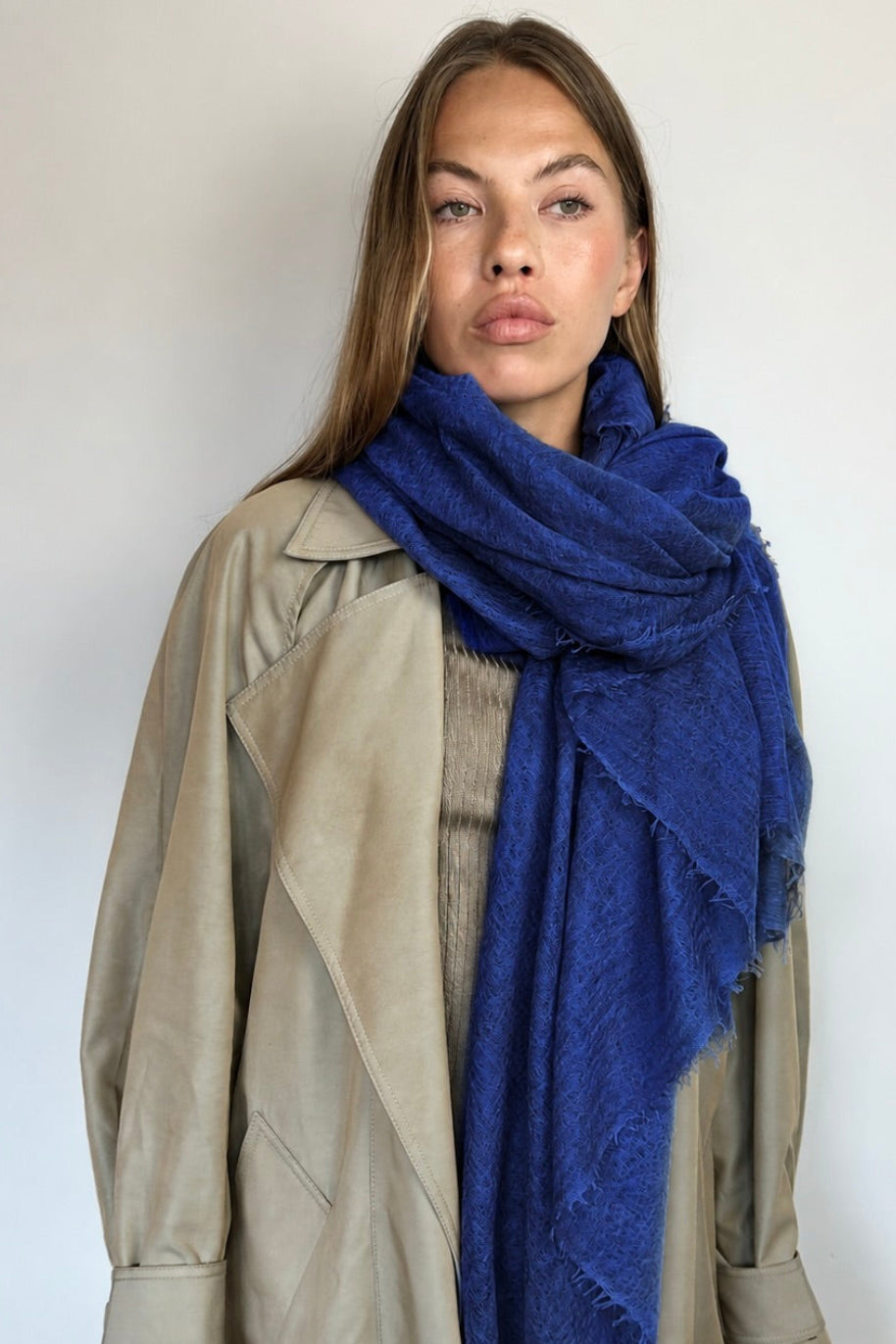 A woman wearing a beige trench coat and a textured royal blue scarf, standing against a plain light gray wall.