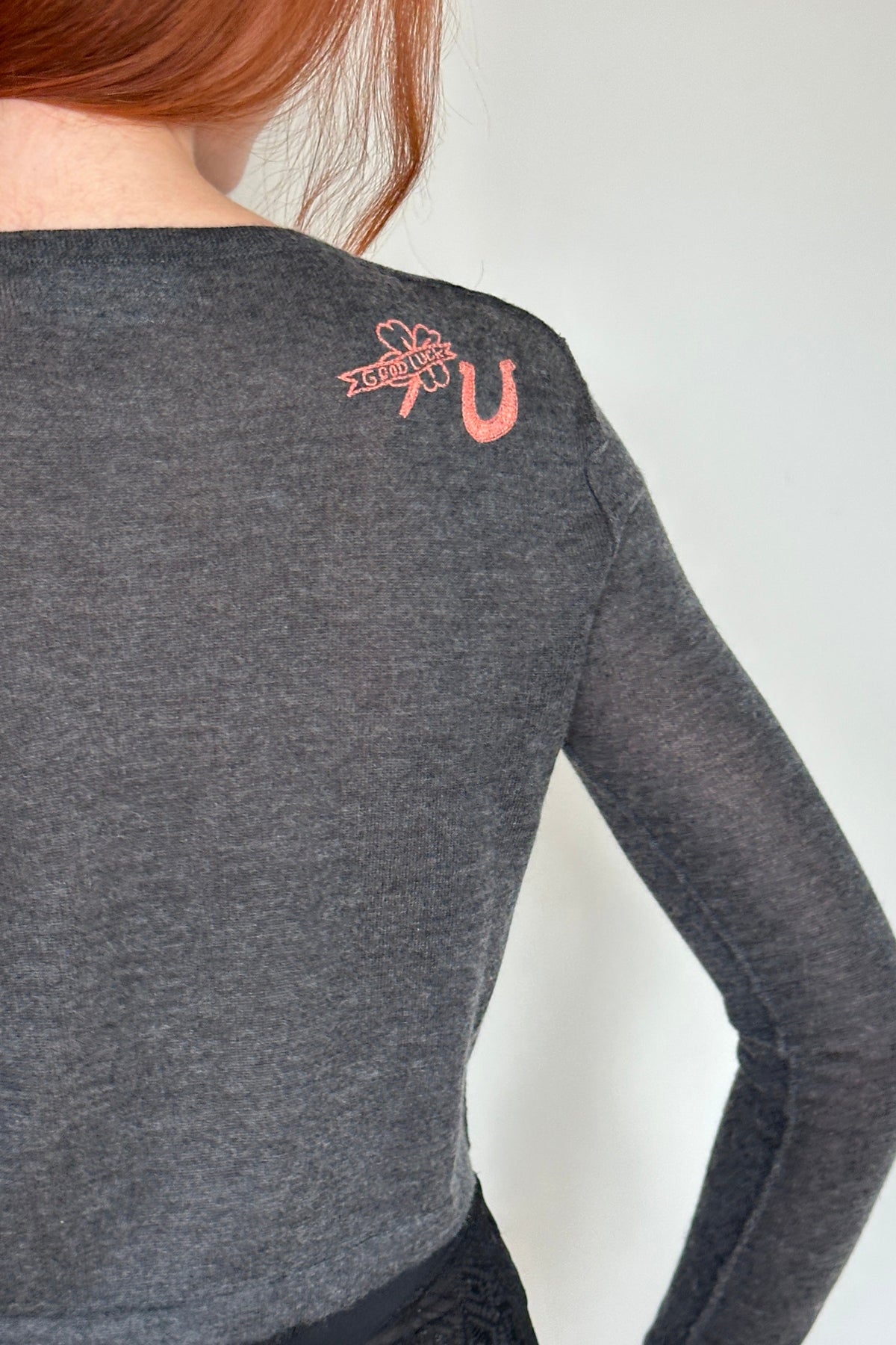 A close-up back view of a person wearing a dark gray long-sleeve crop top with a small coral-pink embroidered graphic on the upper back, paired with a black lace skirt.