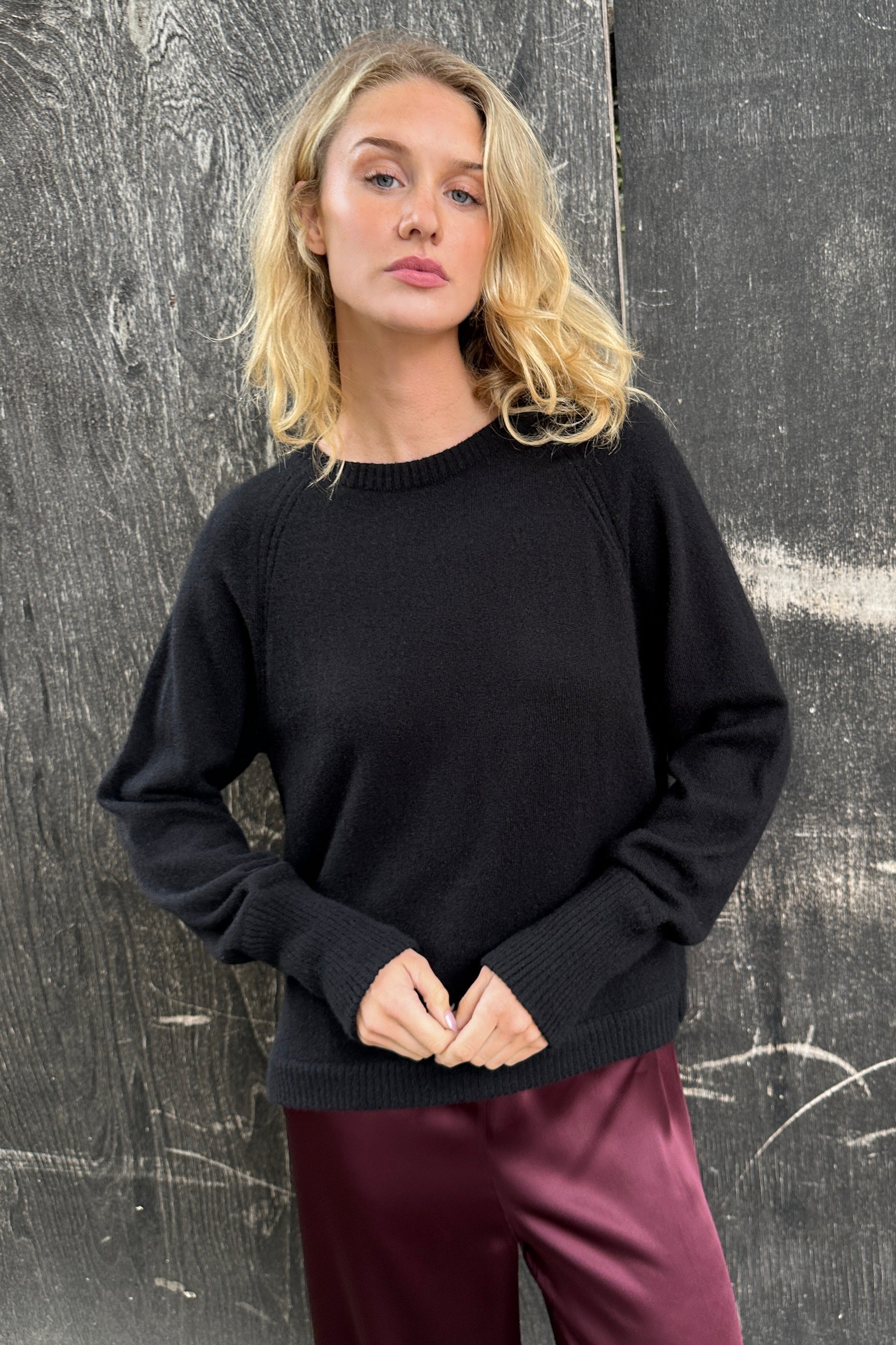 Woman wearing a black cashmere sweater and burgundy pants standing against a textured wall.