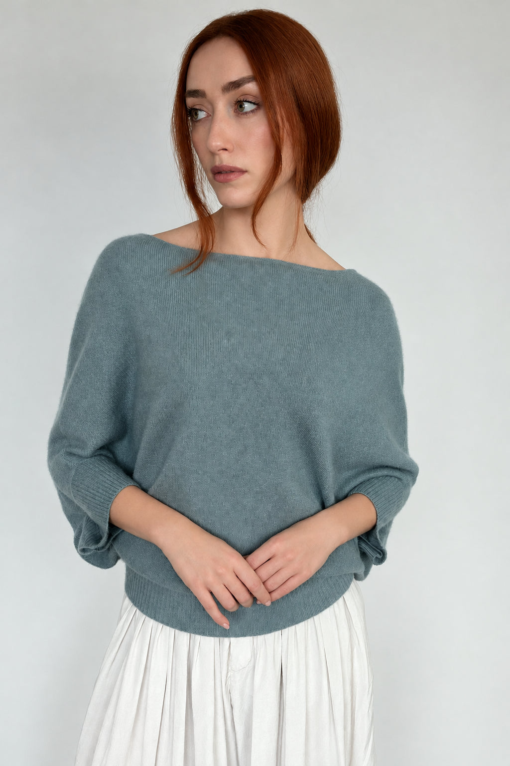 A woman with red hair wearing a muted sage green knit sweater with a wide boat neckline and three-quarter sleeves, paired with a light gray pleated skirt, against a plain light gray background.