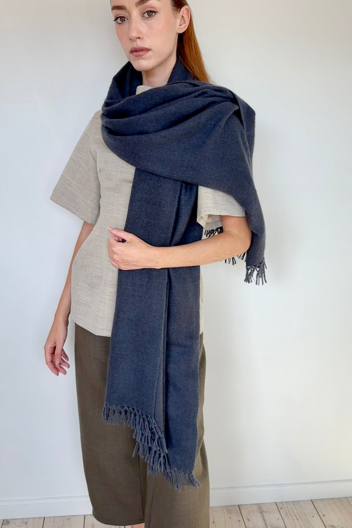 A woman modeling a dark navy blue fringed scarf wrapped around her shoulders, paired with a beige top and brown pants against a white wall and light wood floor.