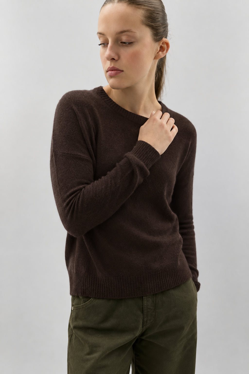 A woman wearing a dark brown long-sleeve knit sweater and olive green pants, standing against a plain light gray background.