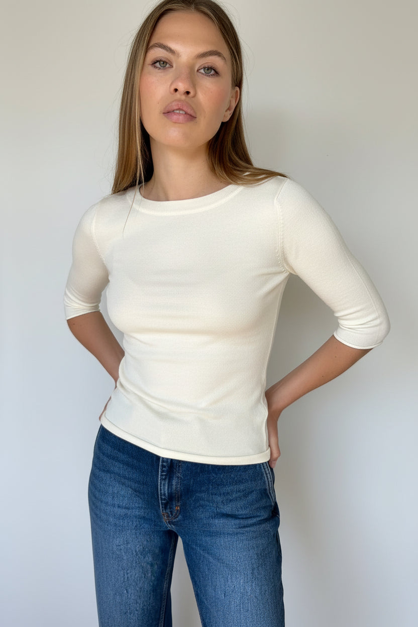 A woman wearing a fitted white short-sleeve top and blue jeans, standing against a plain white wall.