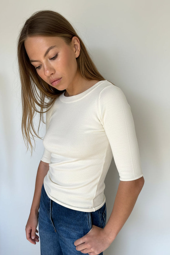 A woman wearing a fittedd whtite short-sleeve top and blue jeans, photographed from above against a plain white wall.