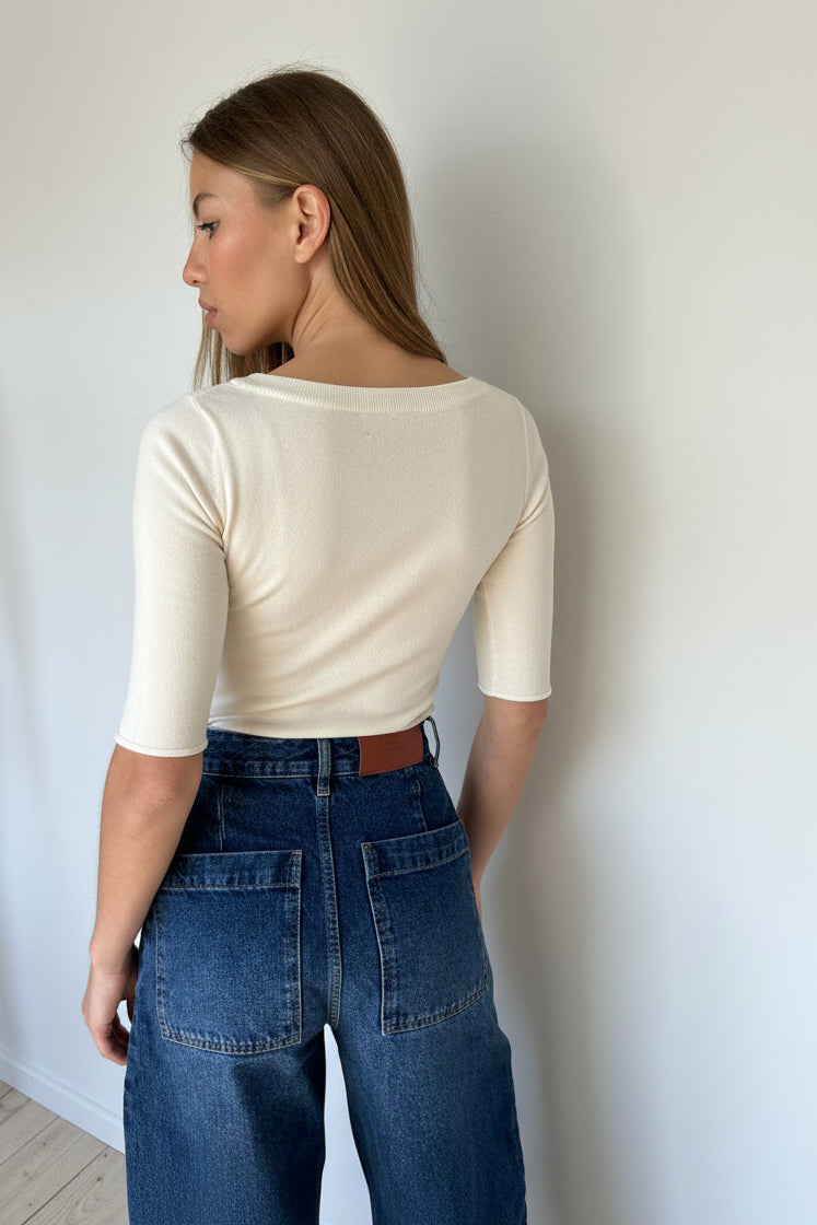 A woman wearing a fitted ivory elbow length sleeve top and blue jeans, photographed indoors against a plain light wall.
