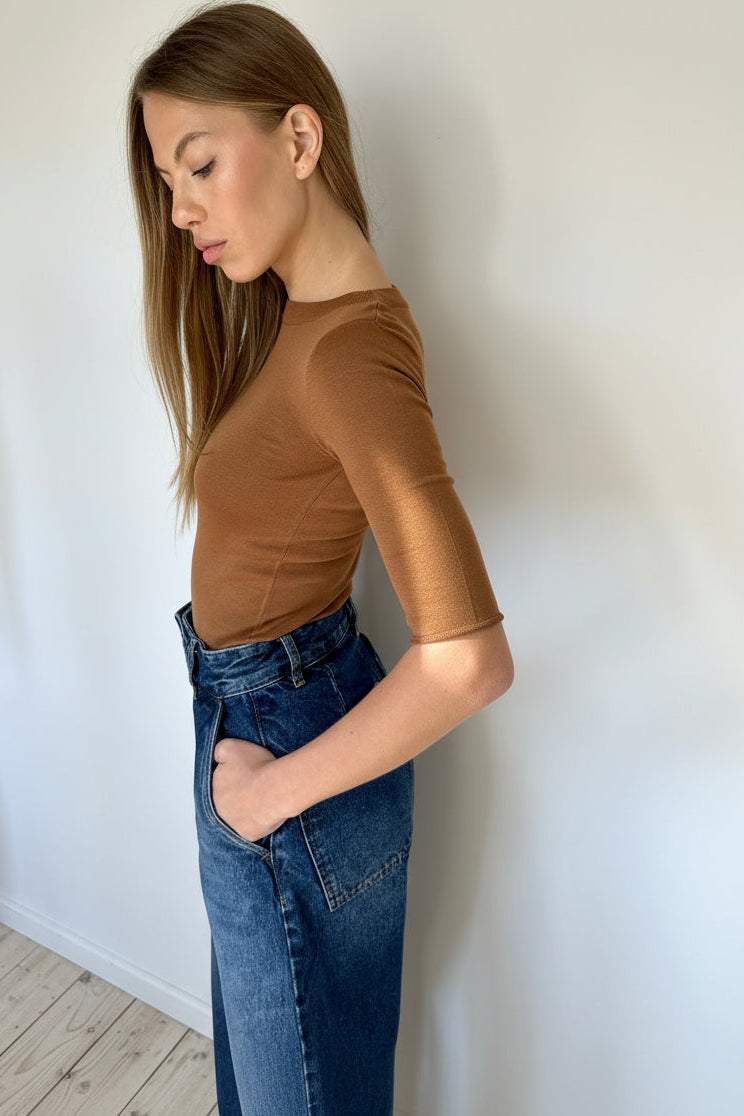 Woman wearing a brown top and blue jeans against a white wall.