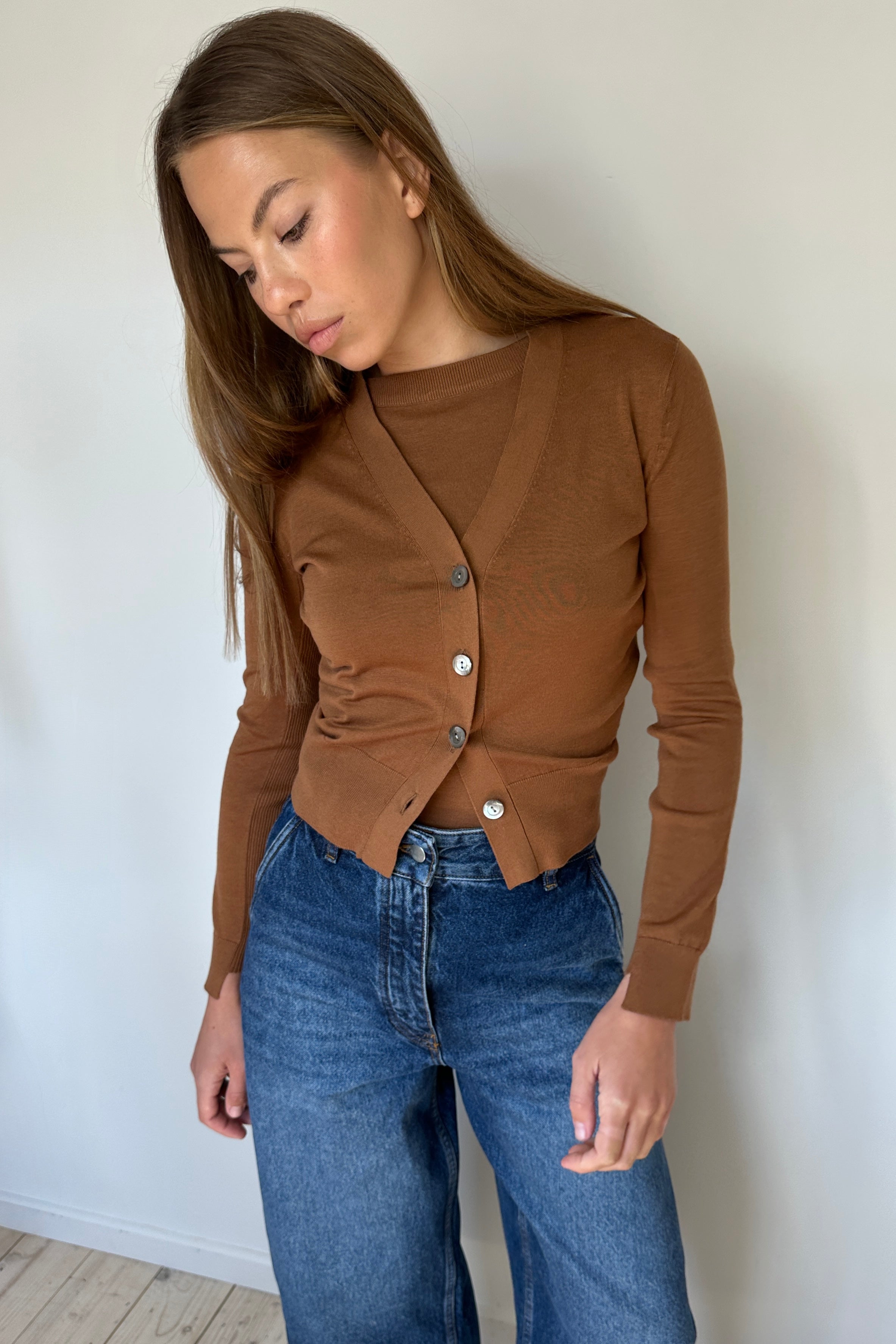 Woman wearing a brown cardigan and blue jeans against a white wall