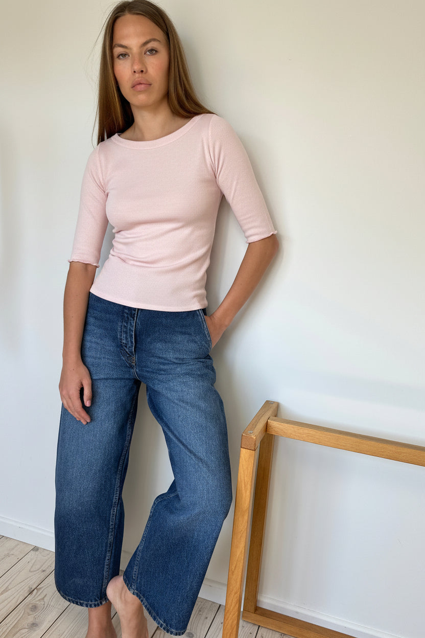 A woman stands barefoot against a white wall wearing a pale pink short-sleeve top and wide-leg blue jeans, with a light wood chair visible on the right.