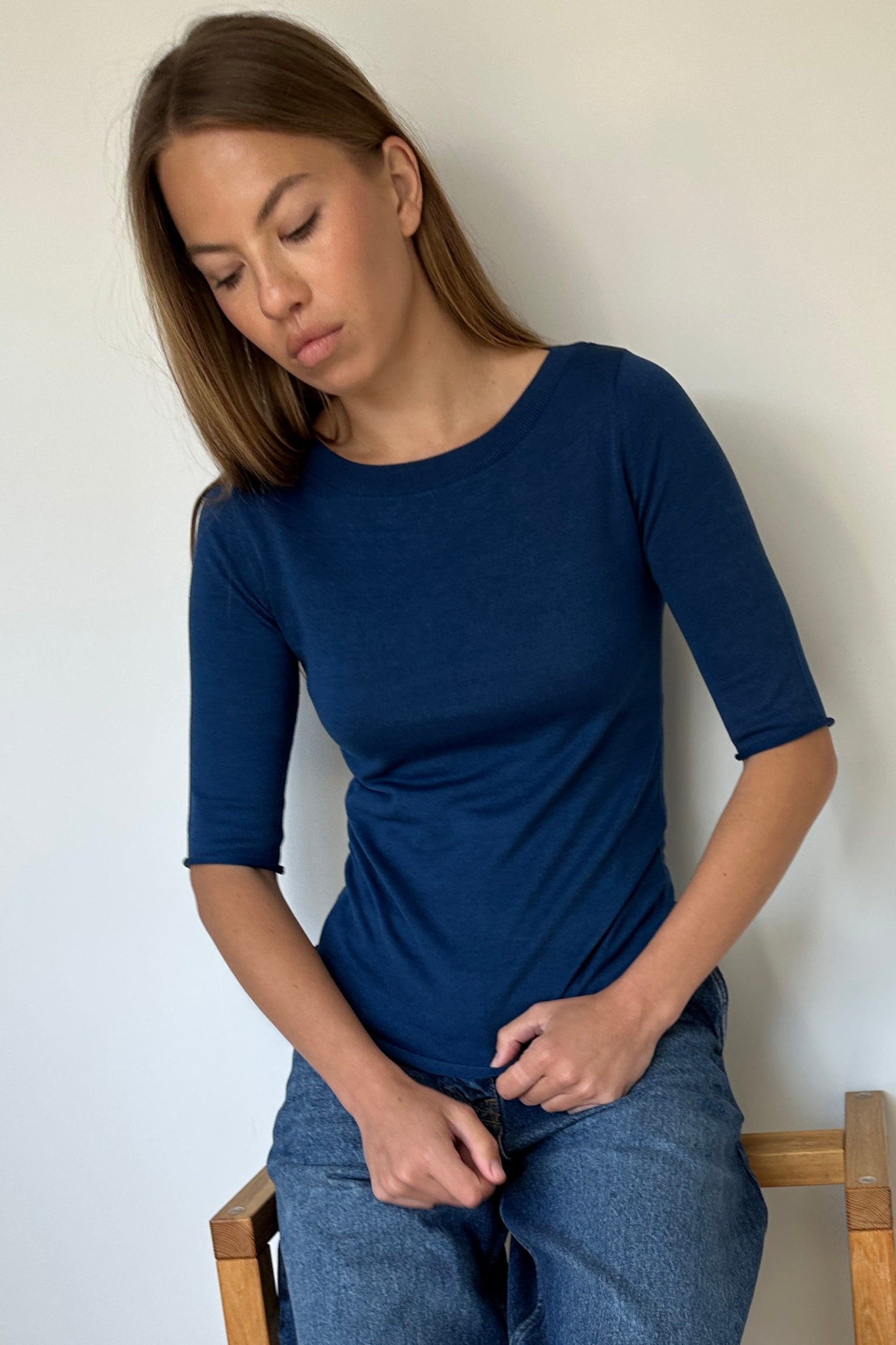 Woman wearing a blue t-shirt and jeans sitting on a chair against a plain wall.