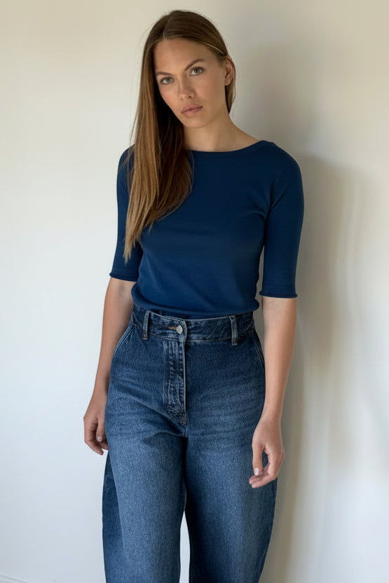 Woman wearing a blue top and jeans standing against a white wall.