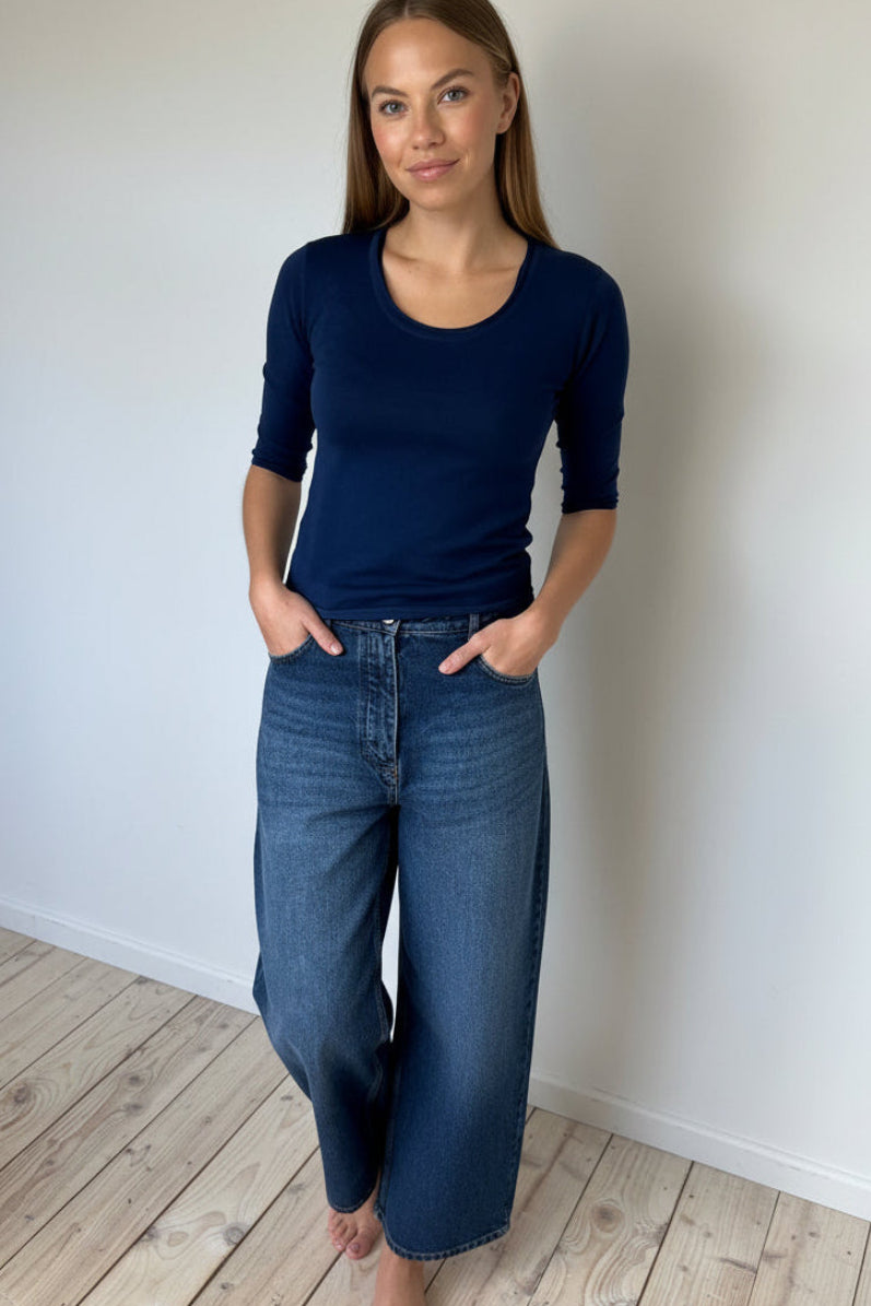 Woman wearing a navy blue top and jeans standing against a white wall.