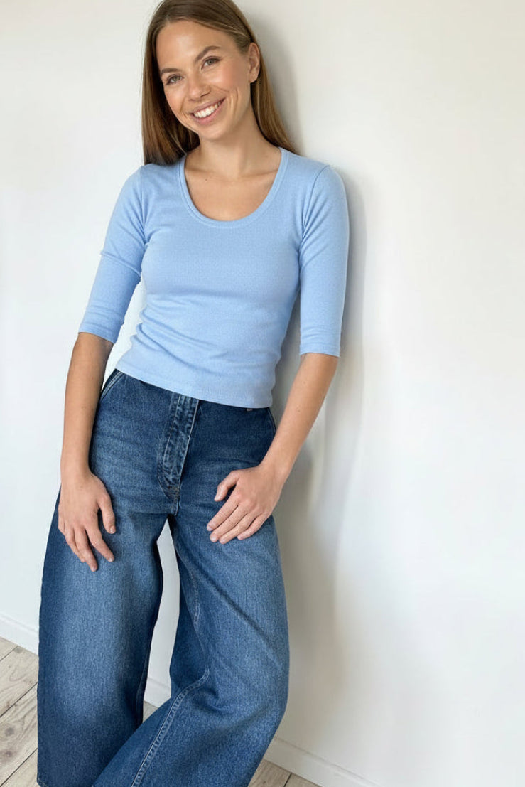 Woman wearing a light blue top and wide-leg jeans against a white wall.