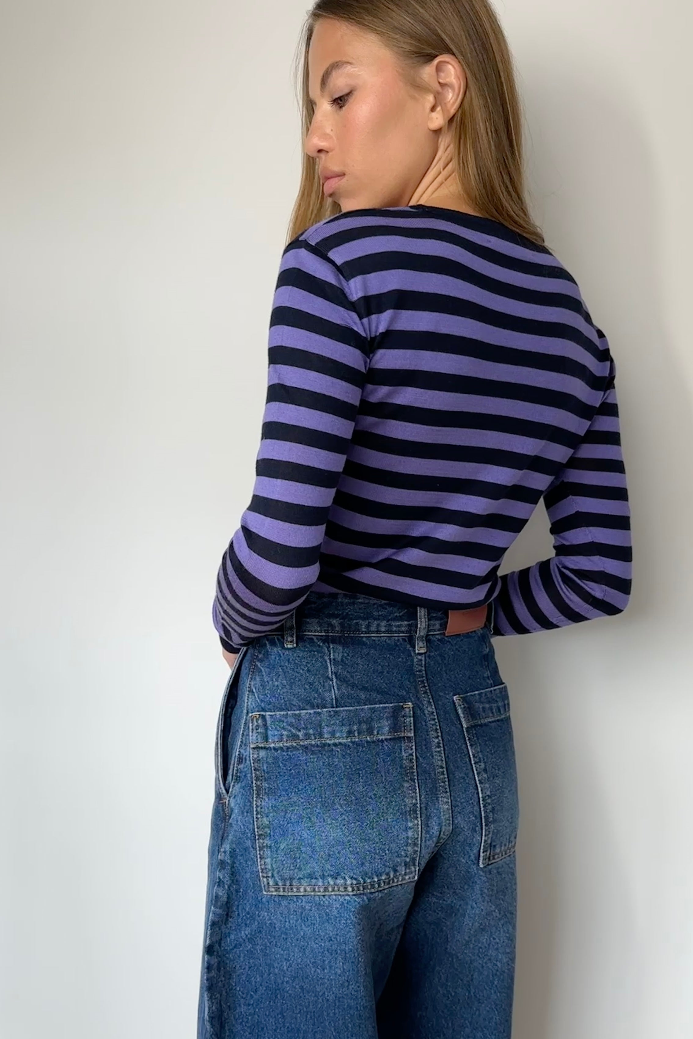 Person wearing a striped long-sleeve shirt and blue jeans against a white wall.