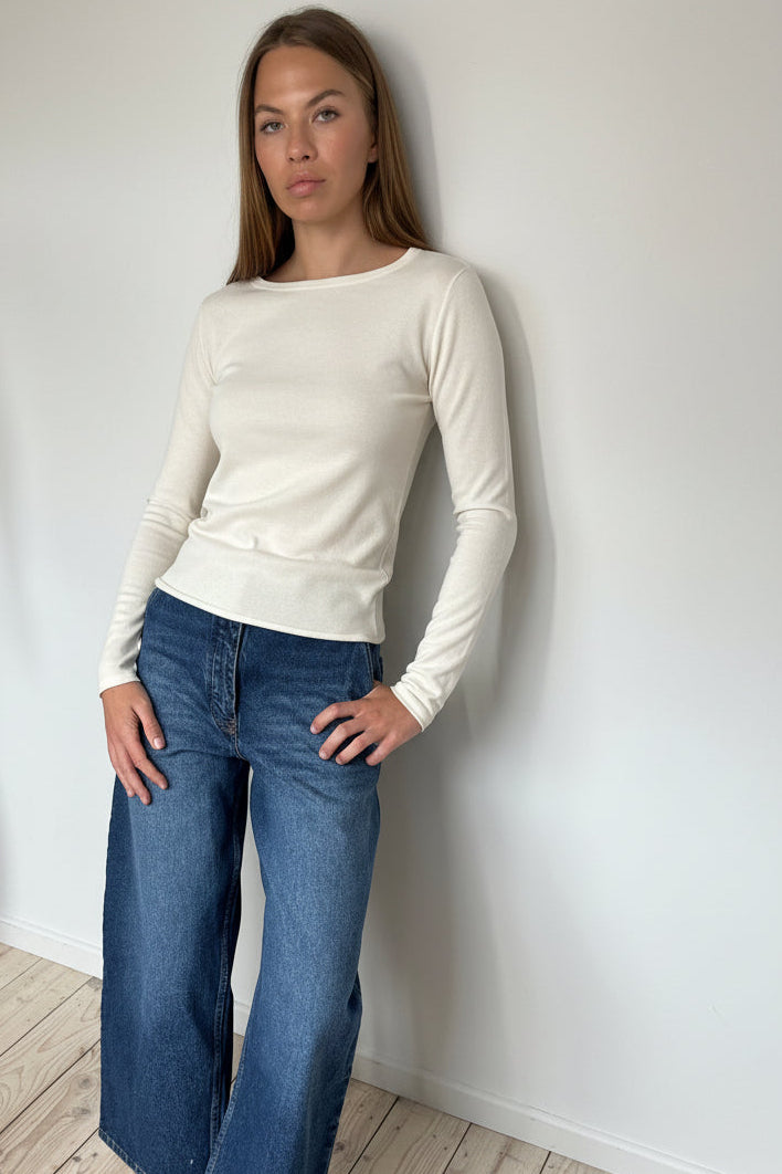 Woman wearing an ivory long-sleeve top and jeans against a white wall.