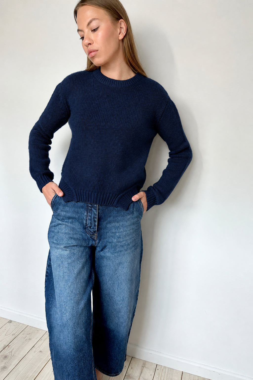 Woman wearing a beige sweater and blue jeans against a white wall.