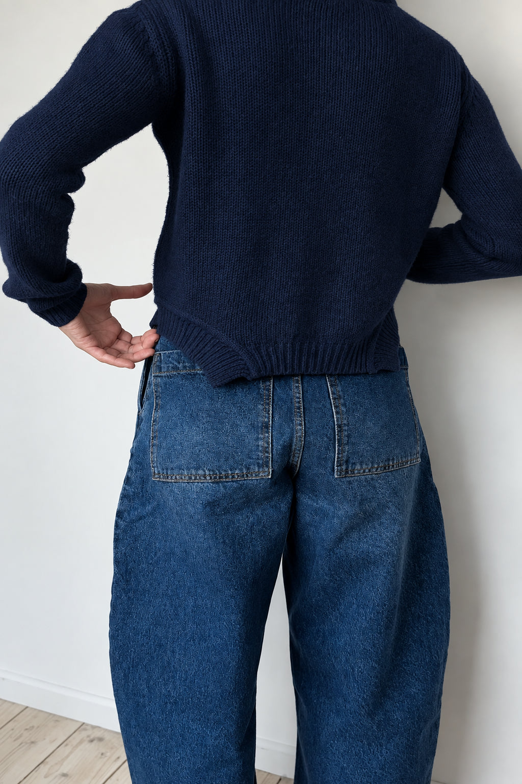 Person wearing a beige sweater and blue jeans against a white wall.