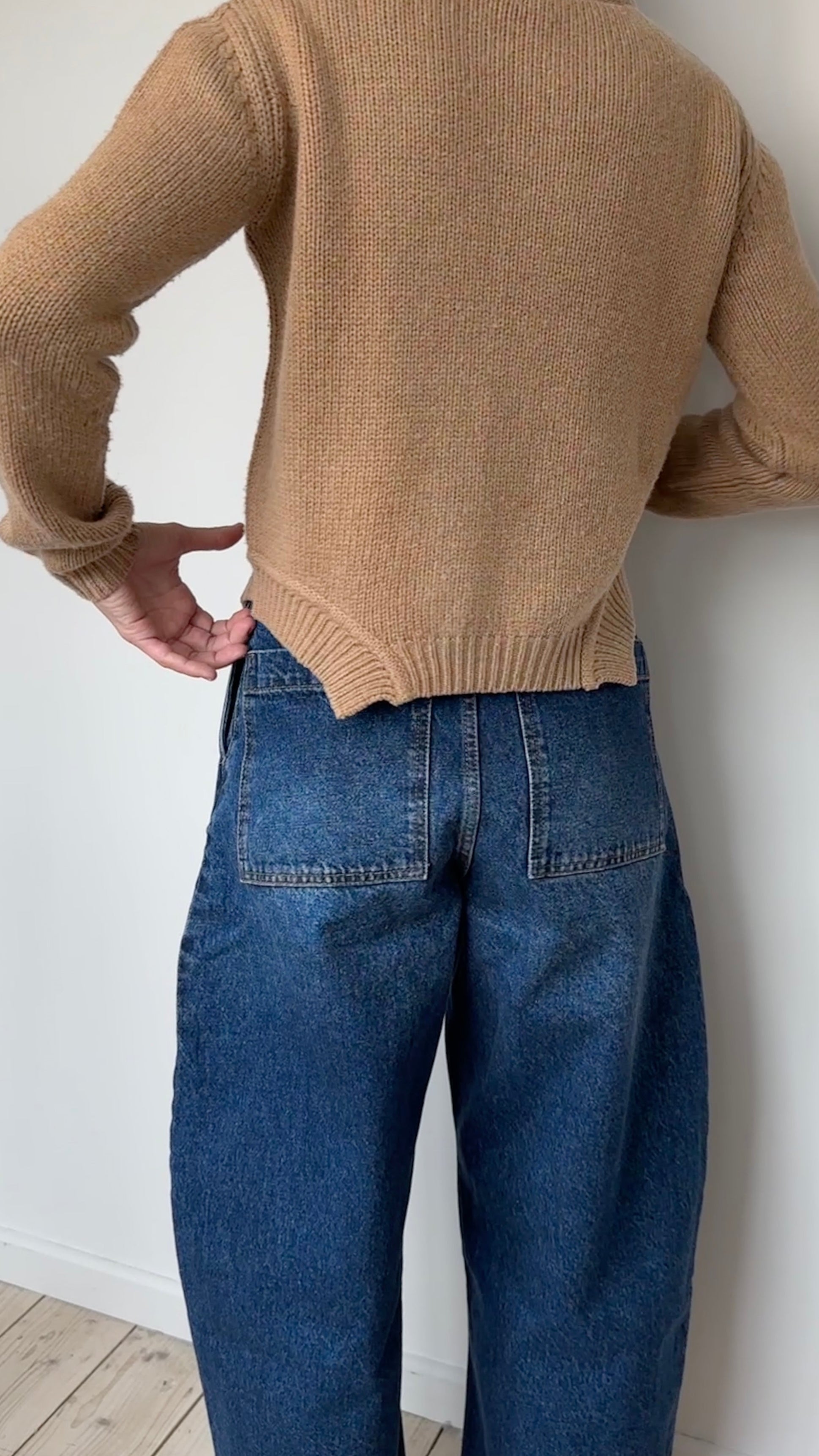 Person wearing a beige sweater and blue jeans against a white wall.