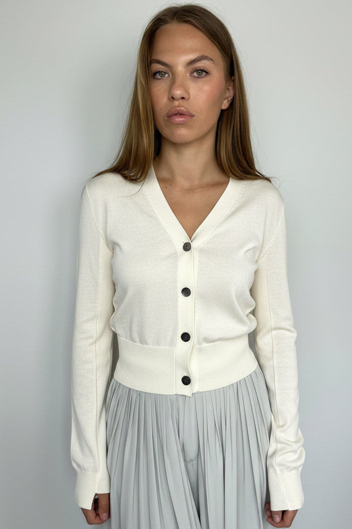 A woman wearing a ivory button-front cardigan and a light gray pleated skirt, photographed against a plain light gray background.